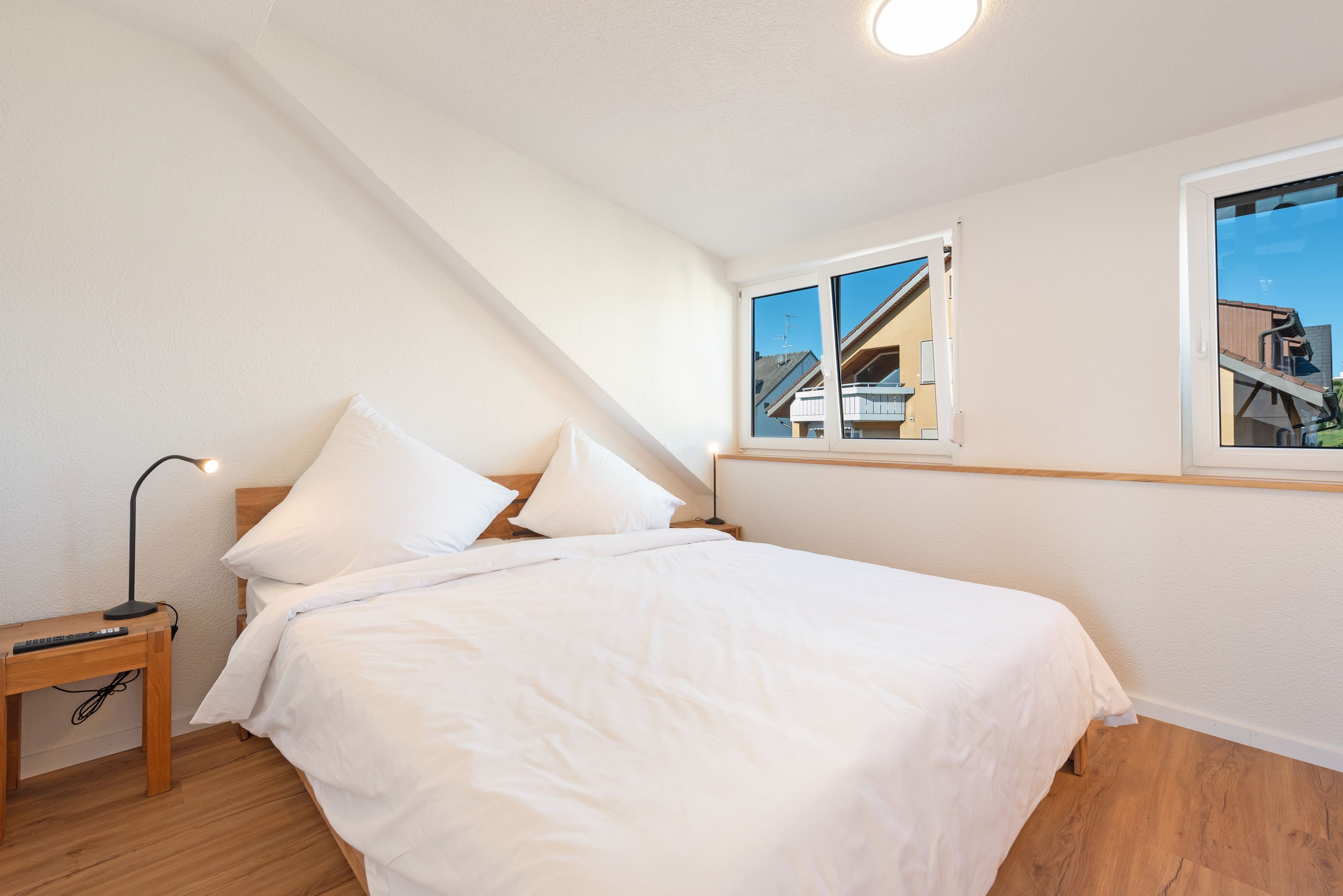 Ein Schlafzimmer mit einem Bett, einem Nachttisch, einer Lampe und einem Fenster mit Blick auf Gebäude.