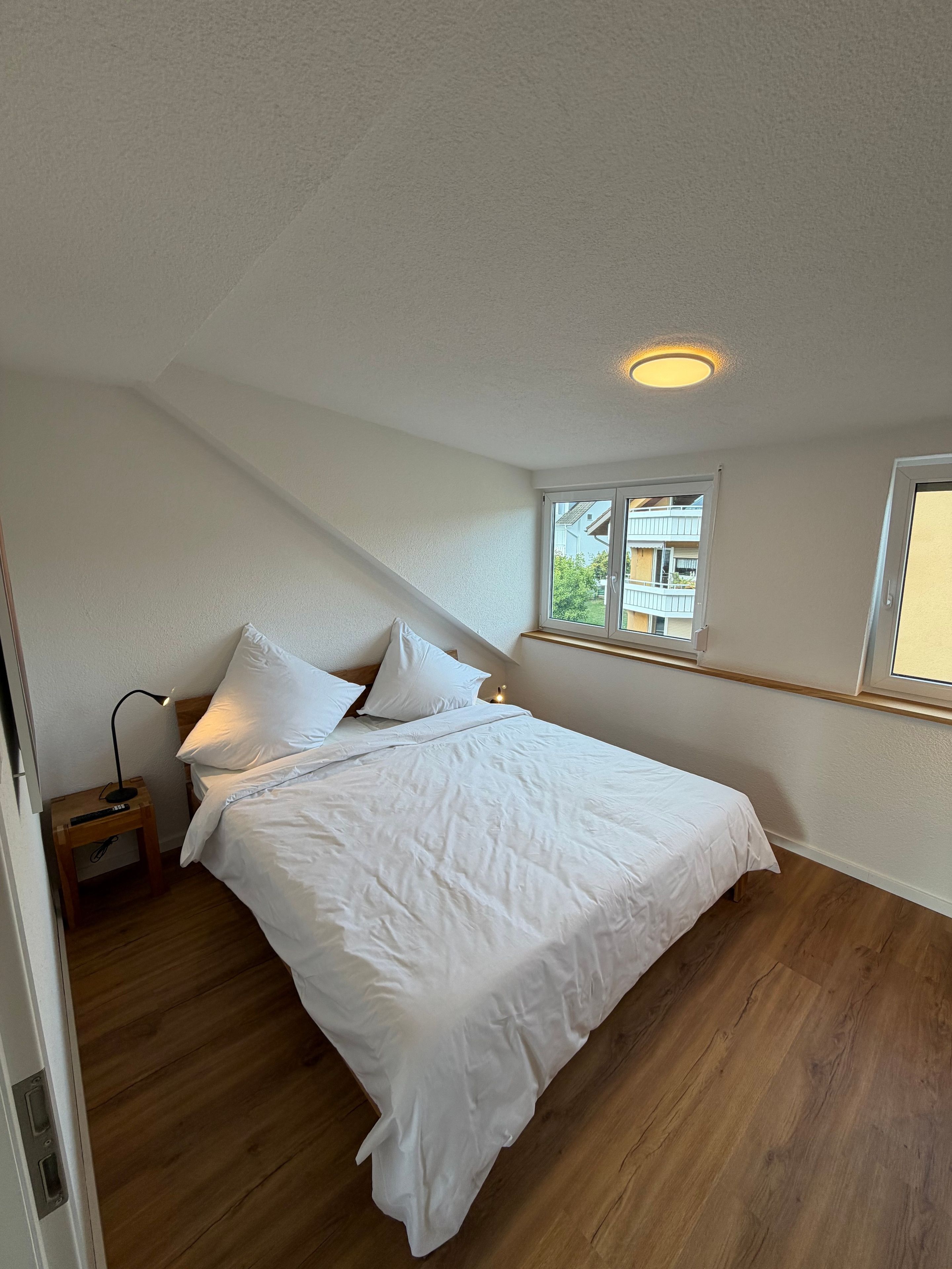 Ein Schlafzimmer mit Bett, Nachttisch, Lampe und einem Fenster mit Blick.