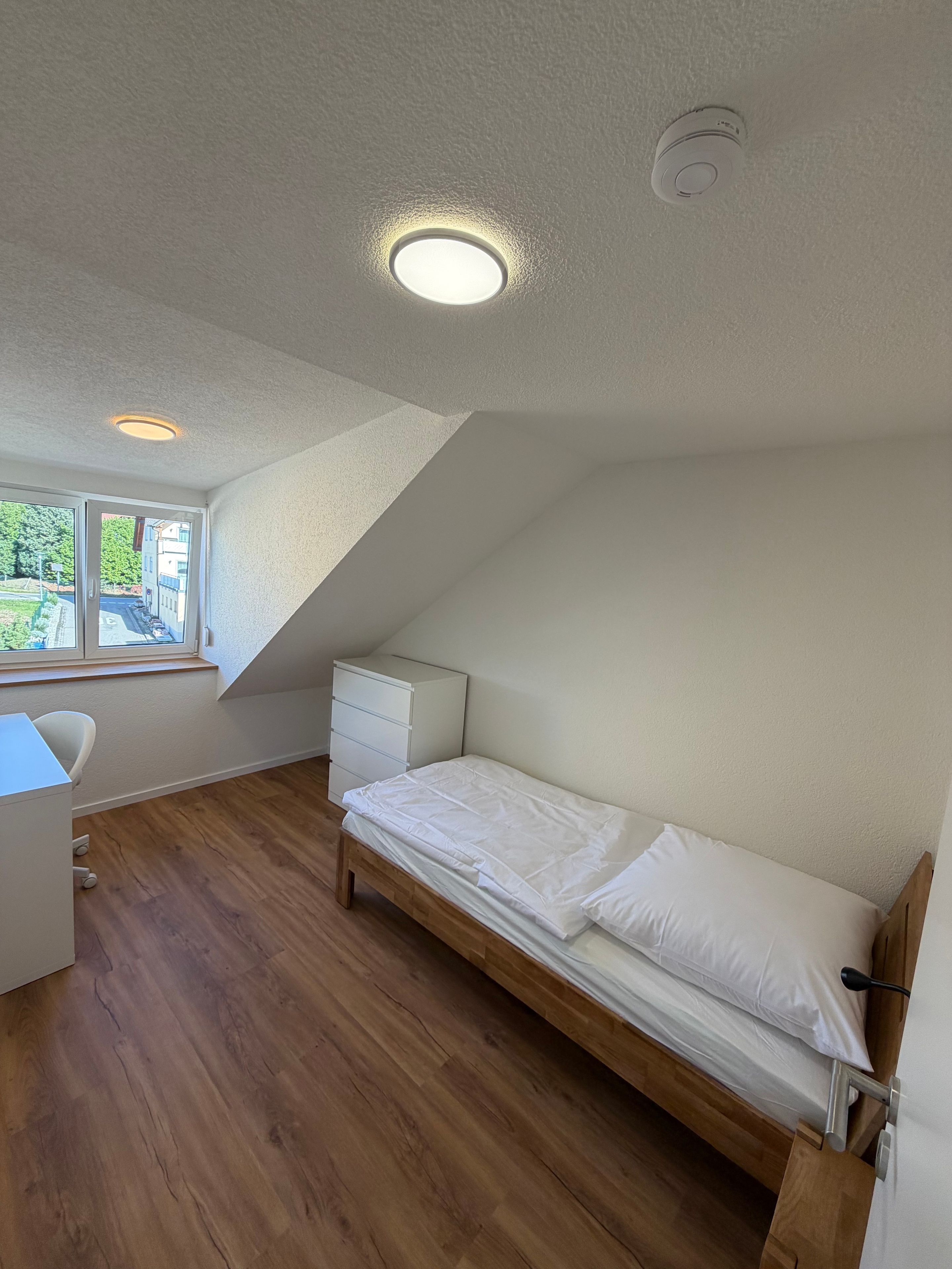 Ein Schlafzimmer mit schrägen Wänden, einem Bett, einem Schreibtisch und einem Fenster.