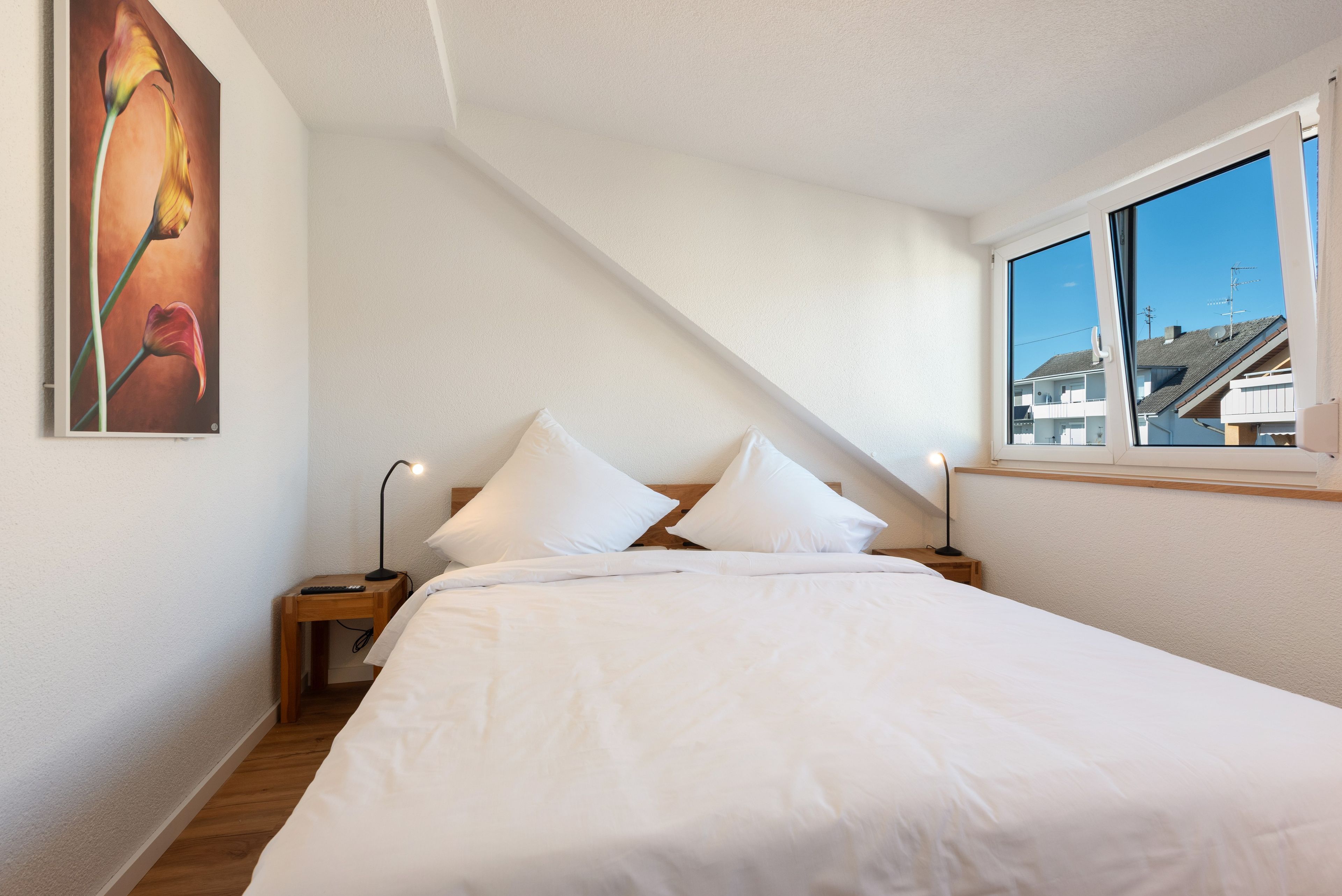 Ein Schlafzimmer mit einem Bett, zwei Nachttischen, einem Schreibtischchen, einem Gemälde und einem Fenster mit Blick auf Gebäude.