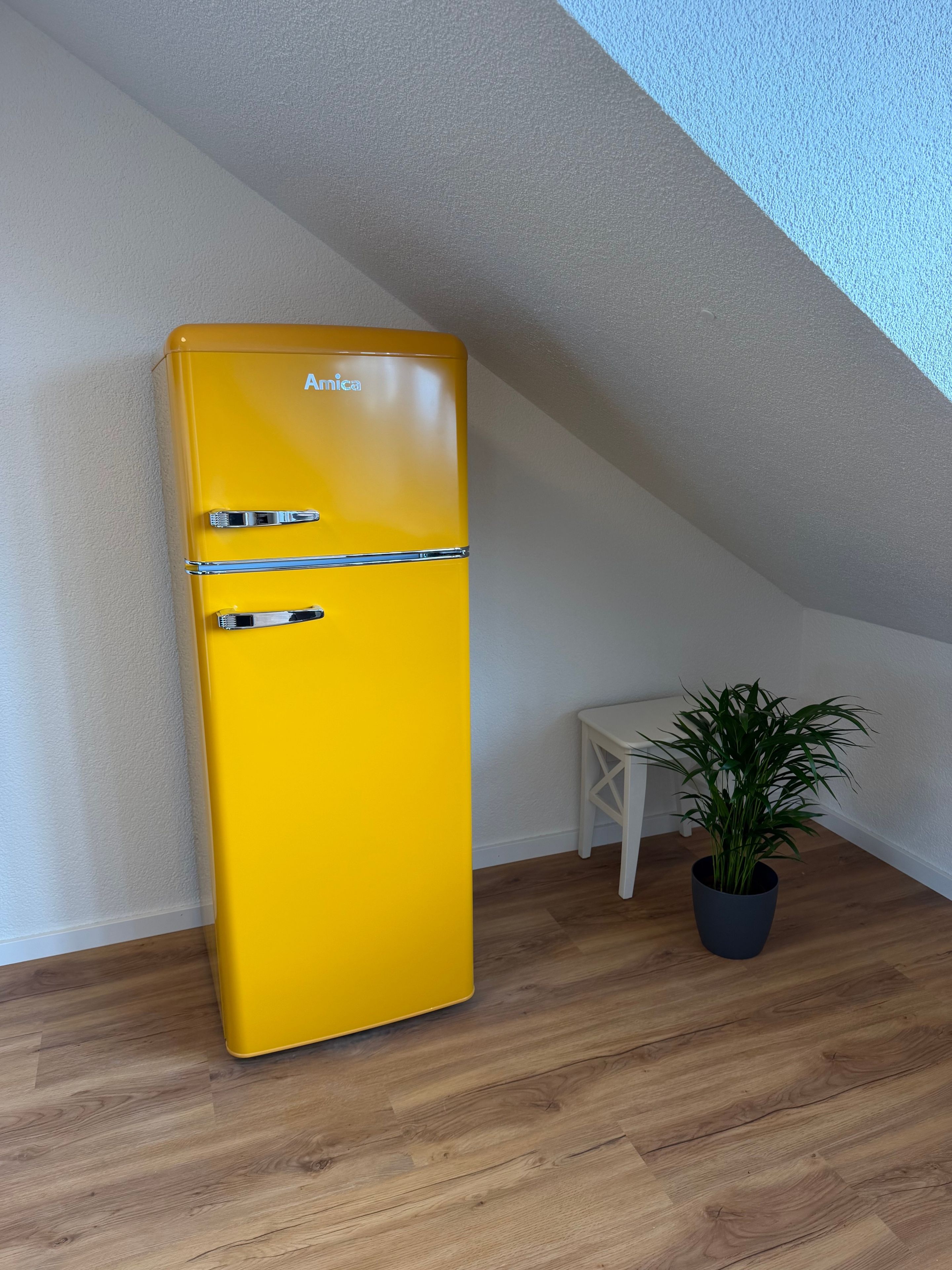 Ein gelber Anica-Kühlschrank steht in einer Ecke neben einem weißen Tisch und einem Topfpflanzen.