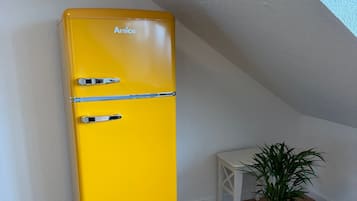 Ein gelber Anica-Kühlschrank steht in einer Ecke neben einem weißen Tisch und einem Topfpflanzen.
