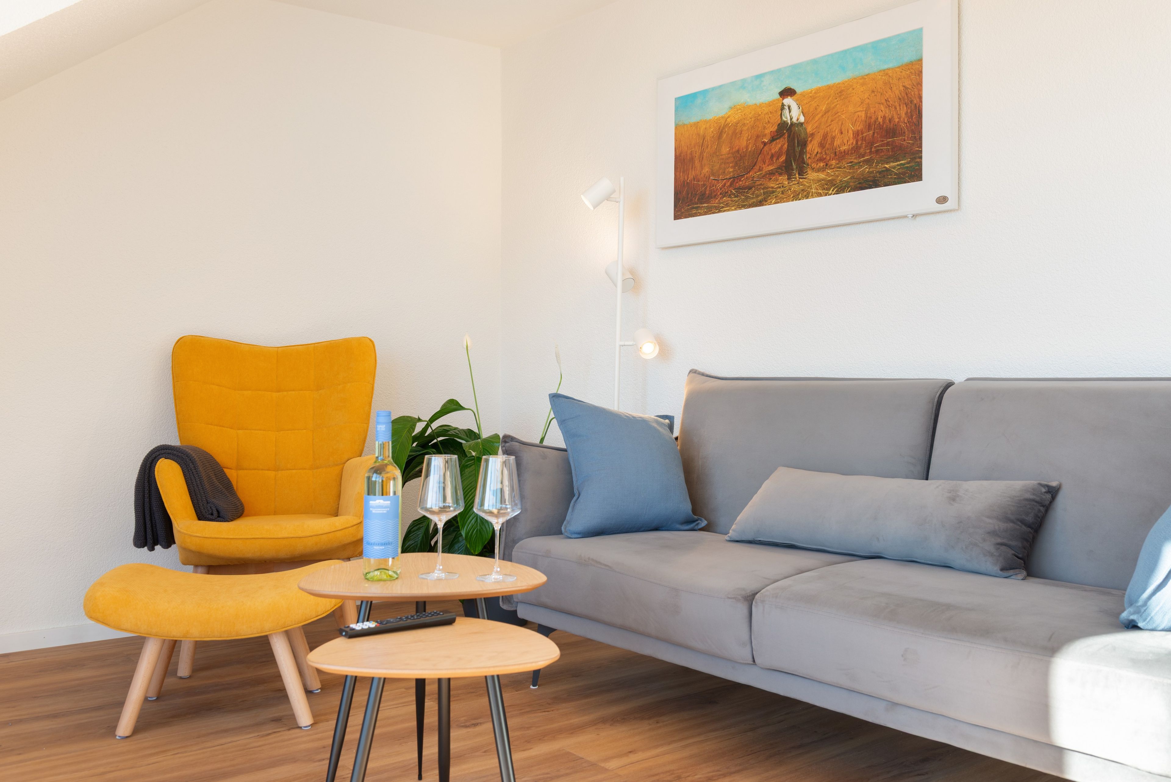 Ein Wohnzimmer mit einer Couch, einem gelben Sessel, einem kleinen runden Tisch mit einer Flasche und Gläsern sowie einem Bild an der Wand.