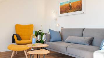 Ein Wohnzimmer mit einer Couch, einem gelben Sessel, einem kleinen runden Tisch mit einer Flasche und Gläsern sowie einem Bild an der Wand.