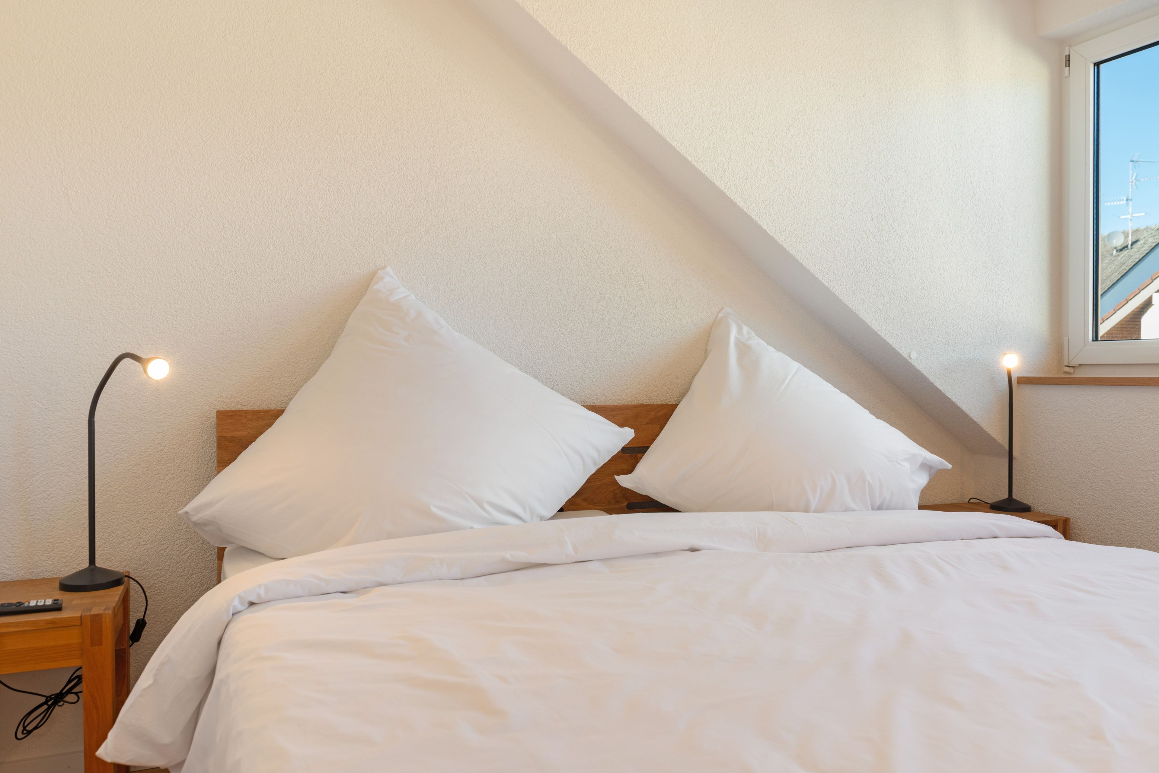 Ein Schlafzimmer mit einem Holz-Kopfteil, zwei weißen Kissen, einer weißen Daunendecke, einem Nachttisch mit einer Lampe und einem Fenster mit Blick zum Himmel.