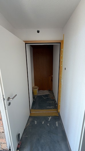 A narrow hallway with a door leading to a room. The hallway has a dark floor, and there is a bucket and a piece of cloth on the floor.