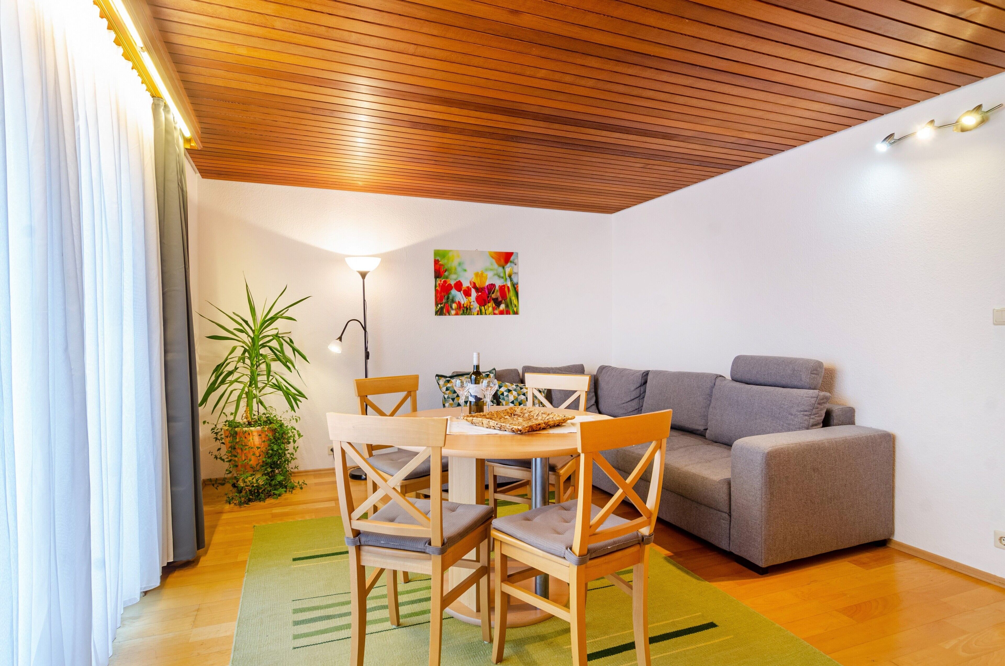Una stanza con soffitto in legno, un tavolo da pranzo con sedie, un divano grigio, una pianta in vaso e un quadro appeso al muro.
