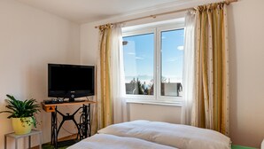 Ein Schlafzimmer mit einem Bett, einem Fernseher auf einem Schrank, einem Fenster mit Vorhängen und einer Zimmerpflanze.