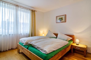 Un dormitorio con una cama de madera, ropa de cama verde, una mesita de noche con una lámpara, una ventana con cortinas translúcidas y un cuadro en la pared.