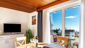 Ein modernes Zimmer mit Flachbildfernseher, Esstisch mit Stühlen, einem Topf mit Pflanze und einem Balkon mit Blick.