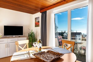 Una habitación moderna con televisor de pantalla plana, una mesa de comedor con sillas, una planta en maceta y un balcón con vistas.