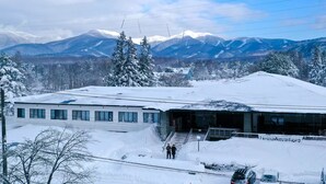 A modern building with a flat roof, large windows, and a covered entrance. Snow covers the ground and trees, with mountains in the background. - LivingAnywhere CommonsAIZU-BANDAI (Bandai)
