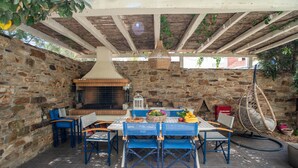 Een terras buiten, met stenen muren, een bakoven, blauwe stoelen, een tafel met fruit en hangmanden.