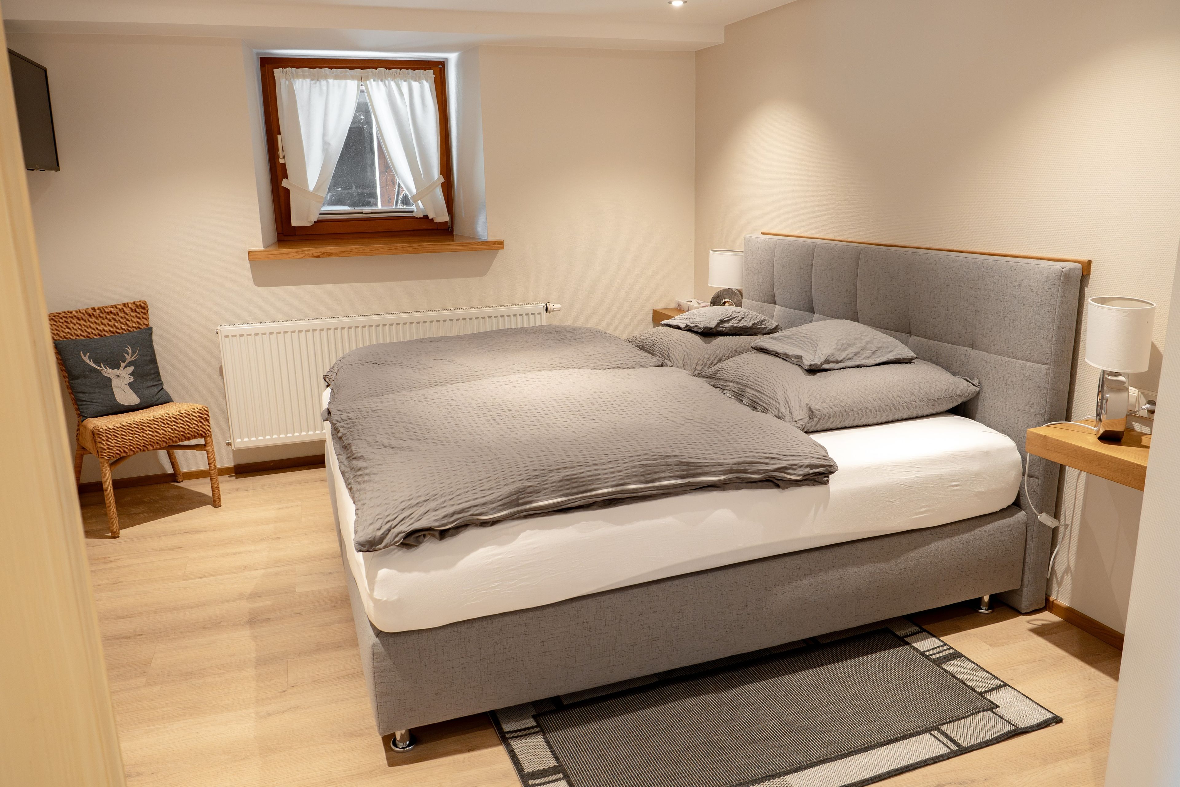 Une chambre à coucher comprenant un lit, une table de chevet, une lampe, une chaise, une fenêtre avec des rideaux et un radiateur.