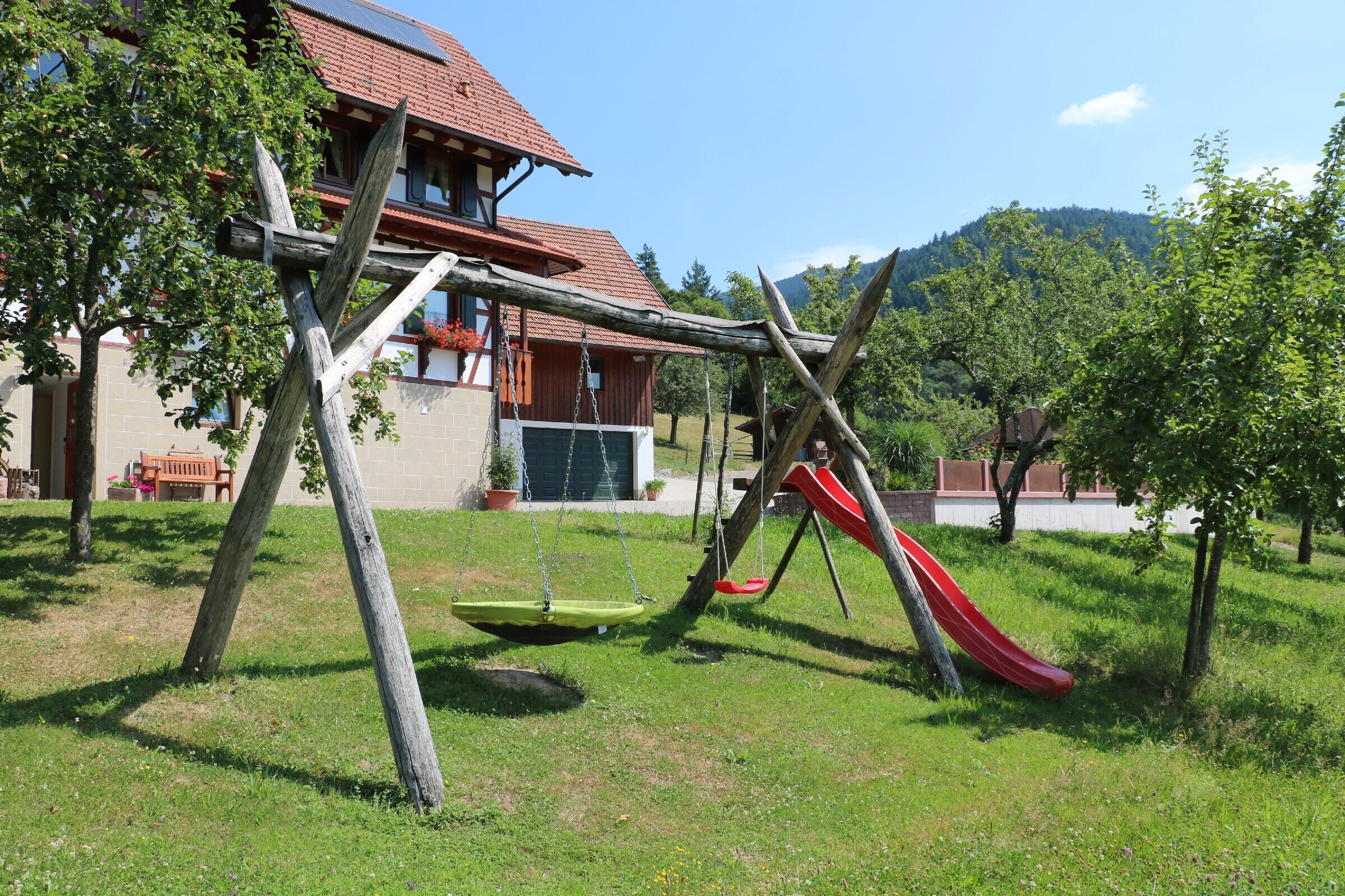 Ein Spielplatz mit Schaukeln und einer Rutsche, ein Gebäude mit rotem Dach und eine begrünte Fläche mit Bäumen.