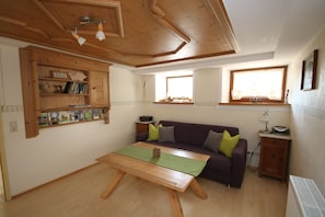 Une pièce avec un plafond en bois, un canapé avec des coussins, une table recouverte d’une nappe verte et une fenêtre avec un rideau.