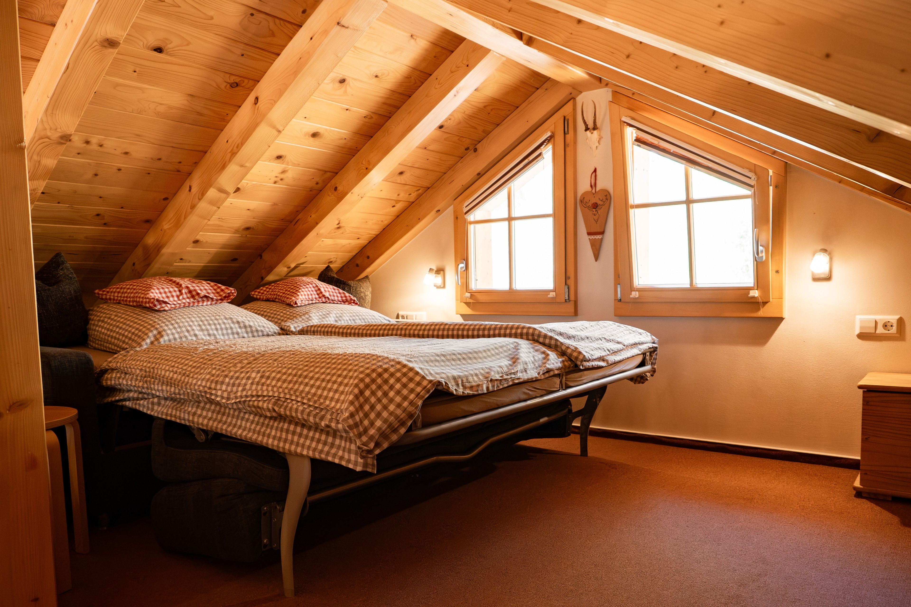 A cozy attic bedroom with a sloped ceiling, two beds with checkered bedding, a window with a decorative heart, and a small wooden dresser.
