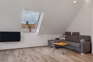 Una sala de estar moderna con un televisor de pantalla plana, un sofá, una mesita de madera y una ventana con vistas a edificios.