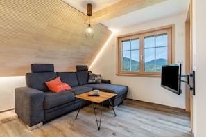 A modern living room with a sofa, a coffee table, a TV mounted on the wall, and a window with a view of the outdoors.
