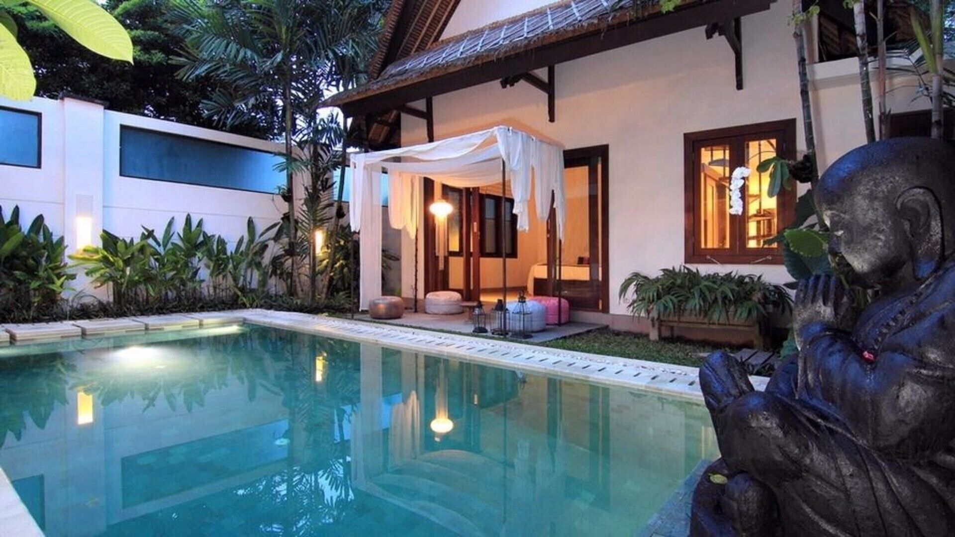 A pool area with a statue, a covered seating area, and a building with windows and plants.