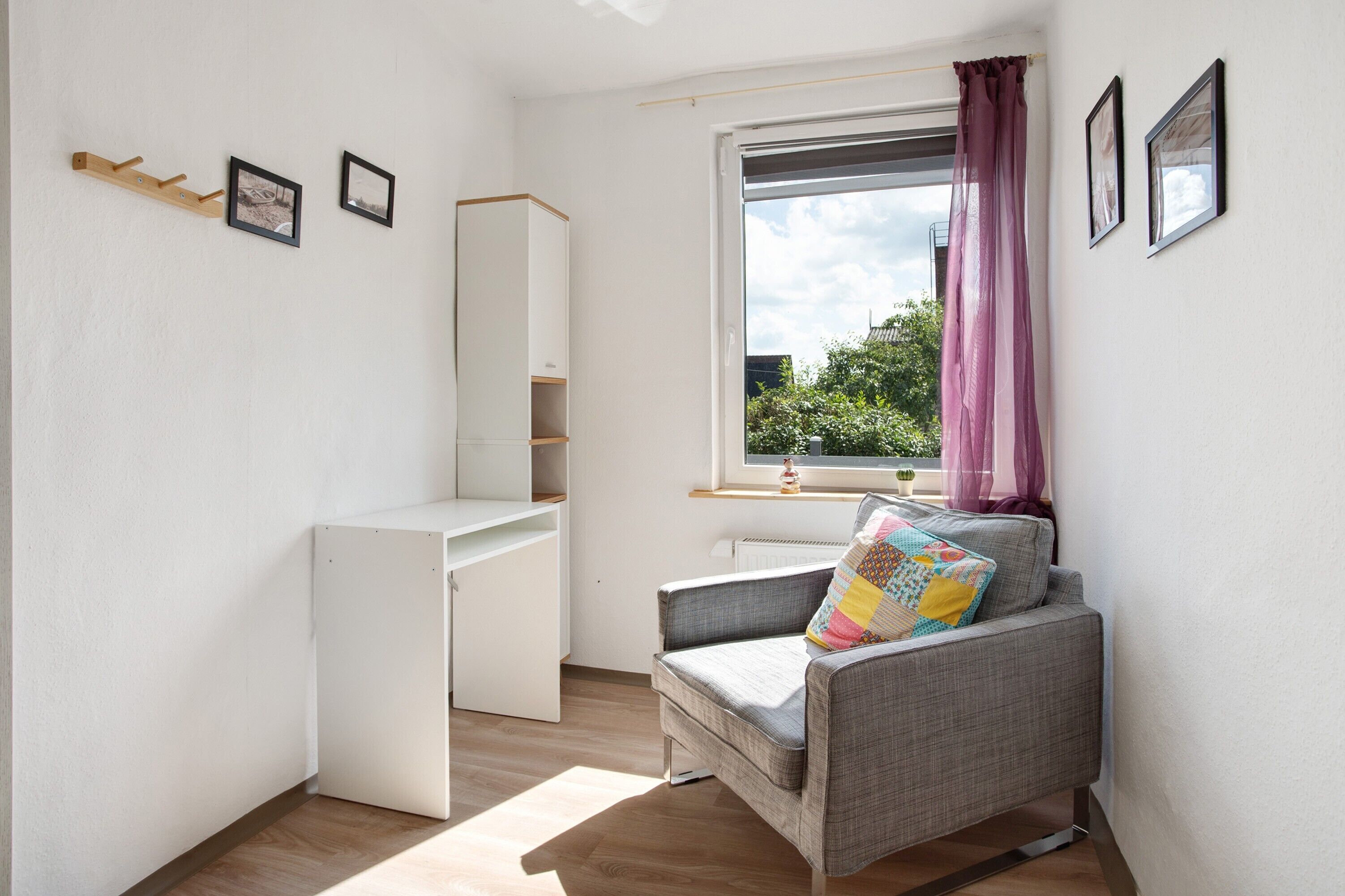 A small, bright room with a desk, chair, and window with curtains.