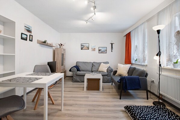 Uma sala de estar moderna com um sofá, uma mesa de jantar e uma mesa de centro. Há prateleiras fixadas na parede e uma televisão.
