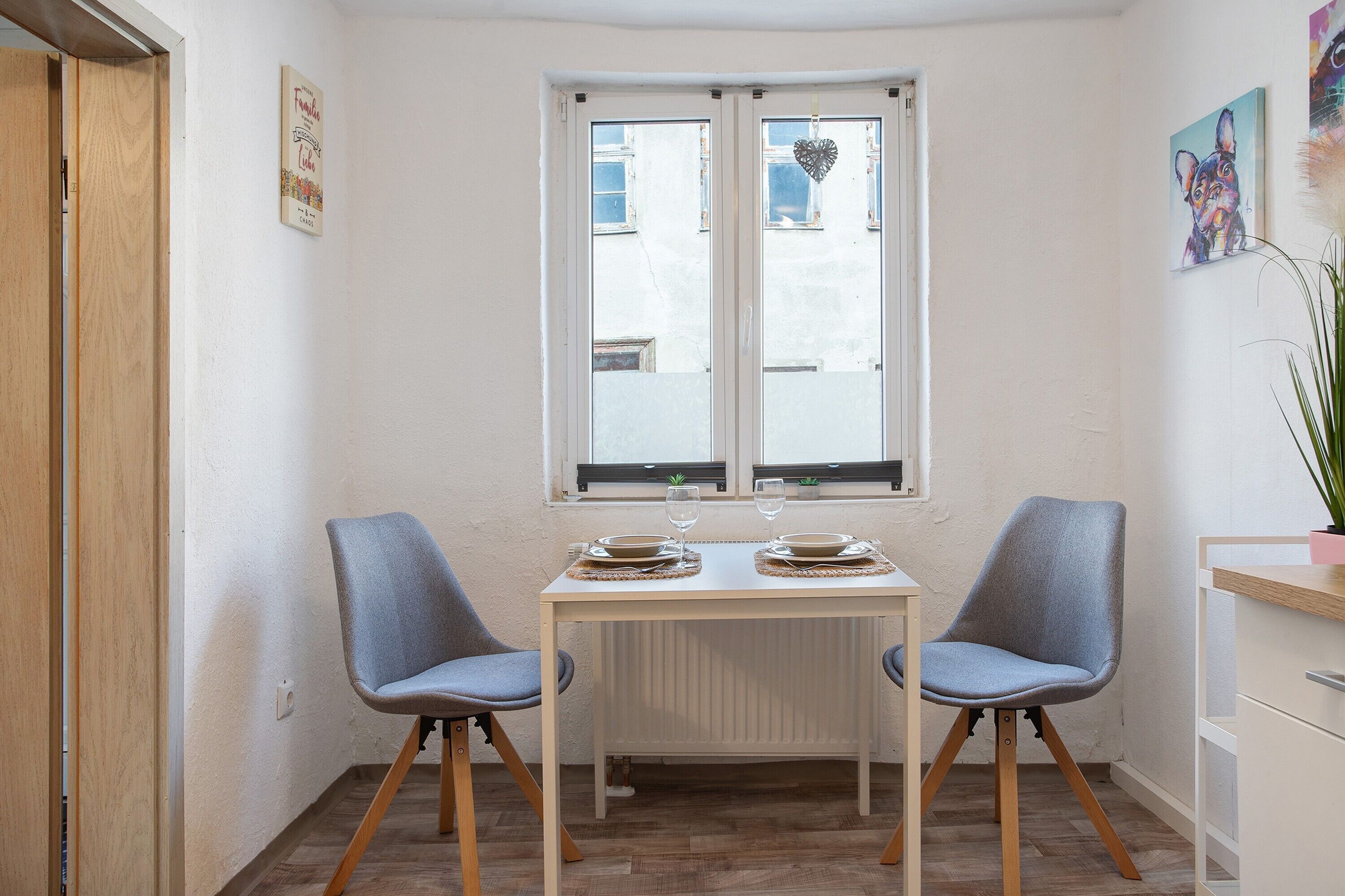 A dining area with a table set for two, two chairs, a window with blinds, and a radiator below the window.