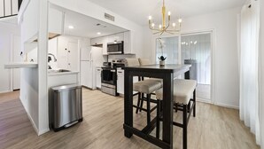 Una cucina moderna con mobili bianchi, un cestino in acciaio inossidabile, un'area pranzo con un tavolo e sedie in legno scuro e un lampadario.