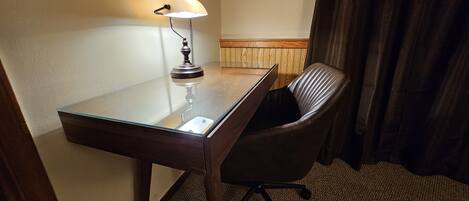 Une chambre d’hôtel avec un bureau en verre, une lampe, un fauteuil en cuir marron et un mur décoratif en bois.