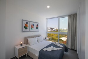 A modern hotel room with a large bed, a nightstand, a wall-mounted artwork, and a view of the city through large windows.