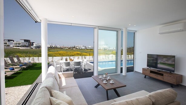 A modern living room with a large sofa, a coffee table, and a TV. There is a balcony with a pool and outdoor seating.