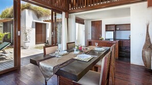 A dining area with a long wooden table set for four, a kitchen in the background, and a view of the outdoors. - Peaceful 16BR Villa W/ Pool in Bali, Bali Villa 2209 (Bali)
