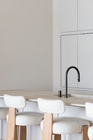A kitchen with a white countertop, a black faucet, and three wooden chairs with white cushions.