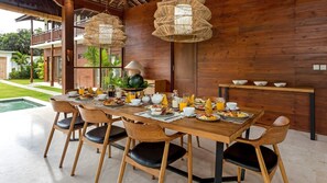 A wooden dining table set for breakfast with chairs, a buffet table, and a view of a garden and building in the background.
