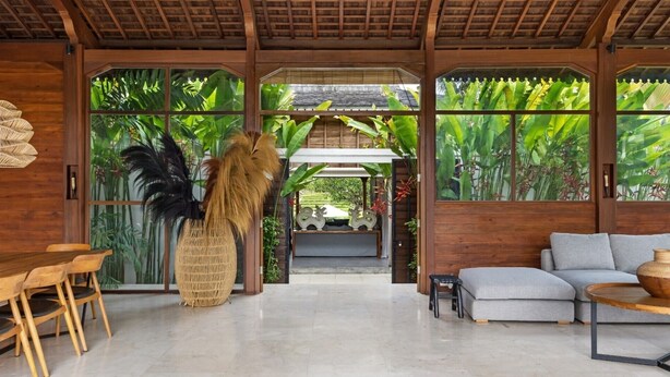 A spacious room with a wooden ceiling, a dining area with wooden chairs, a grey sofa, and a large window with tropical plants.