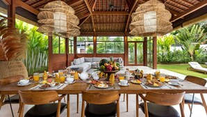 A dining area with a large wooden table set for breakfast, surrounded by chairs, with a view of lush greenery outside.