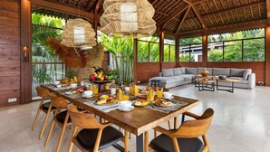 A spacious dining area with a wooden table set for breakfast, surrounded by large windows offering a view of lush greenery.