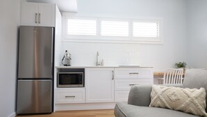 A modern kitchen with white cabinets, a stainless steel refrigerator, a microwave, and a coffee maker. There is a window with blinds above the sink.