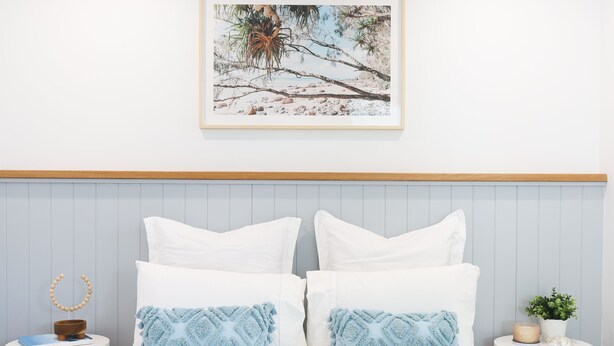 A neatly made bed with white bedding, two decorative pillows, a wooden headboard, and a framed picture above.