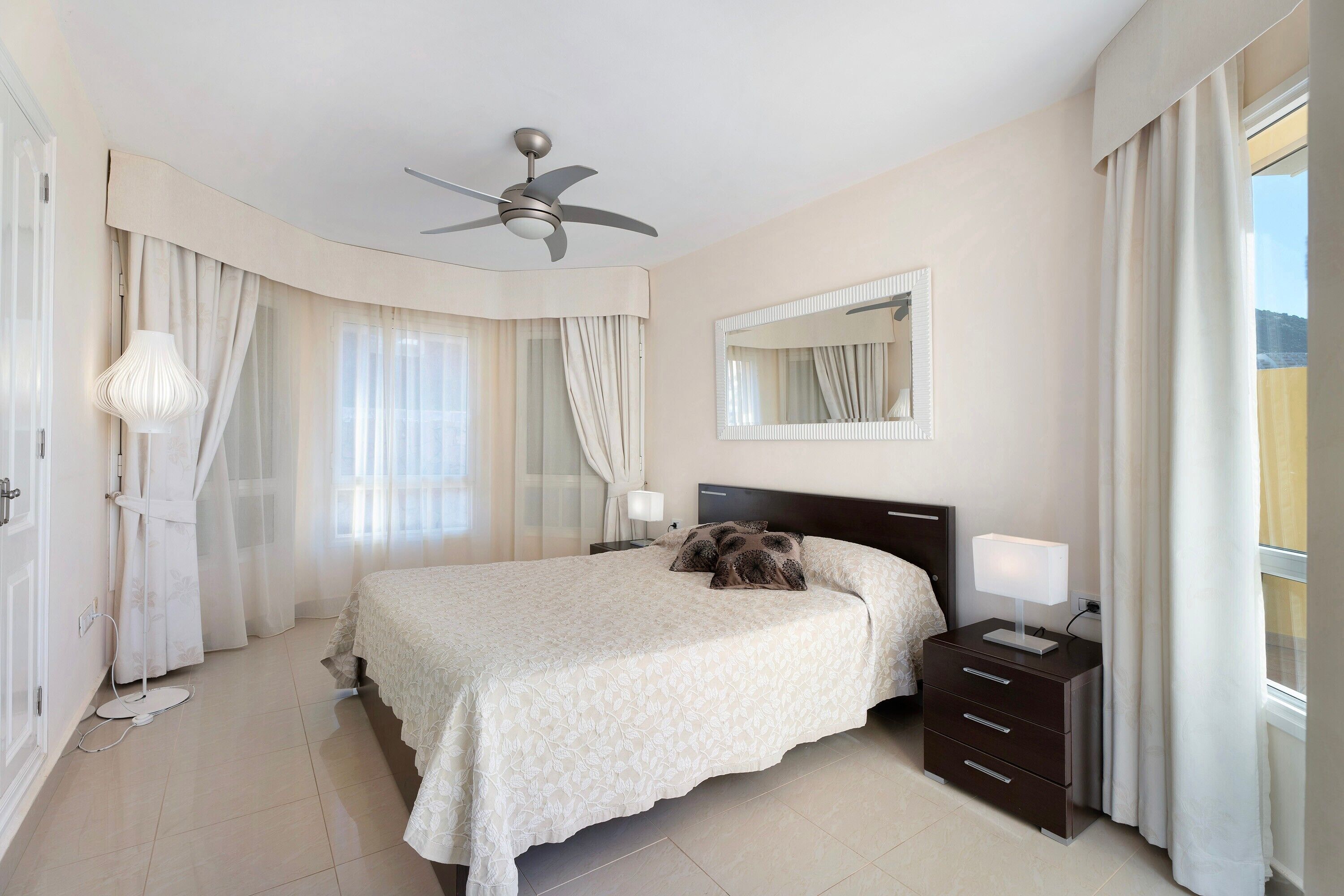 A hotel bedroom with a bed, bedside tables, a ceiling fan, a mirror, and curtains.