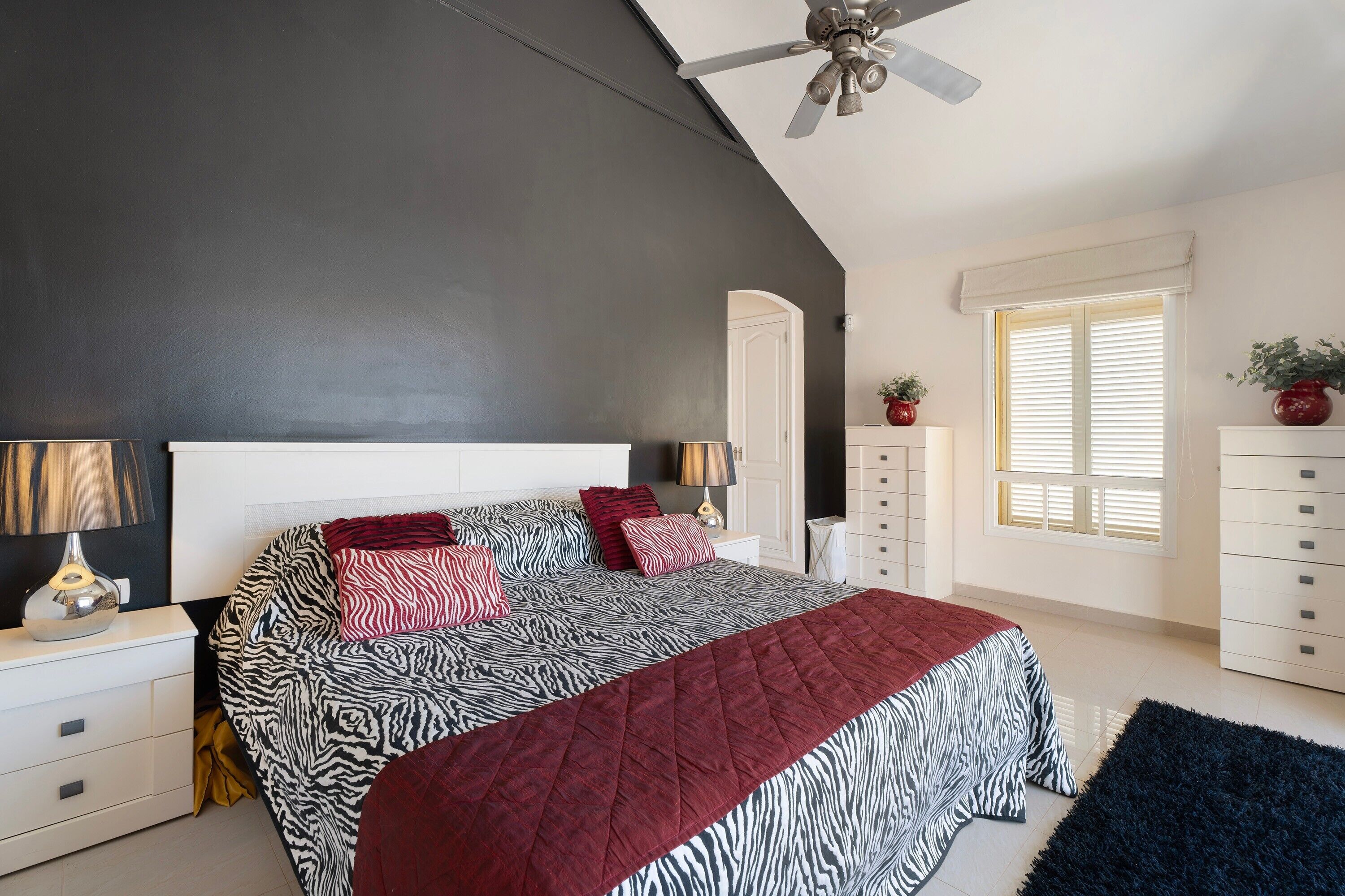 A bedroom with a zebra-patterned bedspread, a red quilt, a ceiling fan, a window with blinds, and a white dresser.
