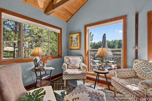 A cozy living room with two sofas, a coffee table, and a side table with a lamp. There are large windows offering a view of trees and a patio.