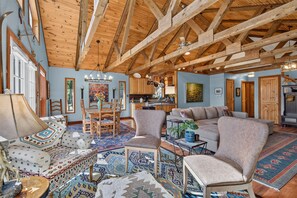 A spacious living room with a high wooden ceiling, a dining area with a wooden table and chairs, and a kitchen in the background.