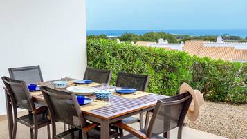 A dining table set outdoors with chairs, a view of the sea, and a hedge in the background.