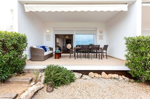 Un espacio exterior cubierto con una mesa y sillas, un sofá de mimbre y vistas a una habitación con una lámpara y un cuadro enmarcado a través de una puerta de vidrio.