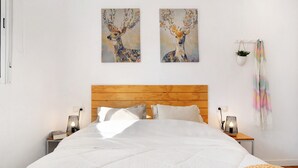 Une chambre à coucher avec une tête de lit en bois, du linge de lit blanc, deux tableaux représentant des cerfs accrochés au mur, des lampes de chevet, une petite plante en pot et un tissu coloré suspendu.