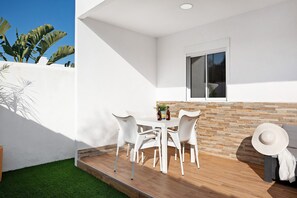 A patio with a table and chairs, a window, and a view of a white wall and plants.