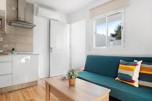A compact living space with a kitchenette, a sofa with colorful cushions, and a wooden table with a plant. - Zahora Qg - Cosy Country House in Barbate, Wi-Fi & A/C (Barbate)