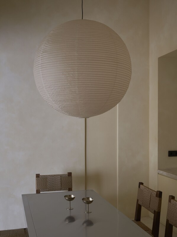 A dining area with a round, textured hanging light fixture, a rectangular table, and two chairs with woven backs.