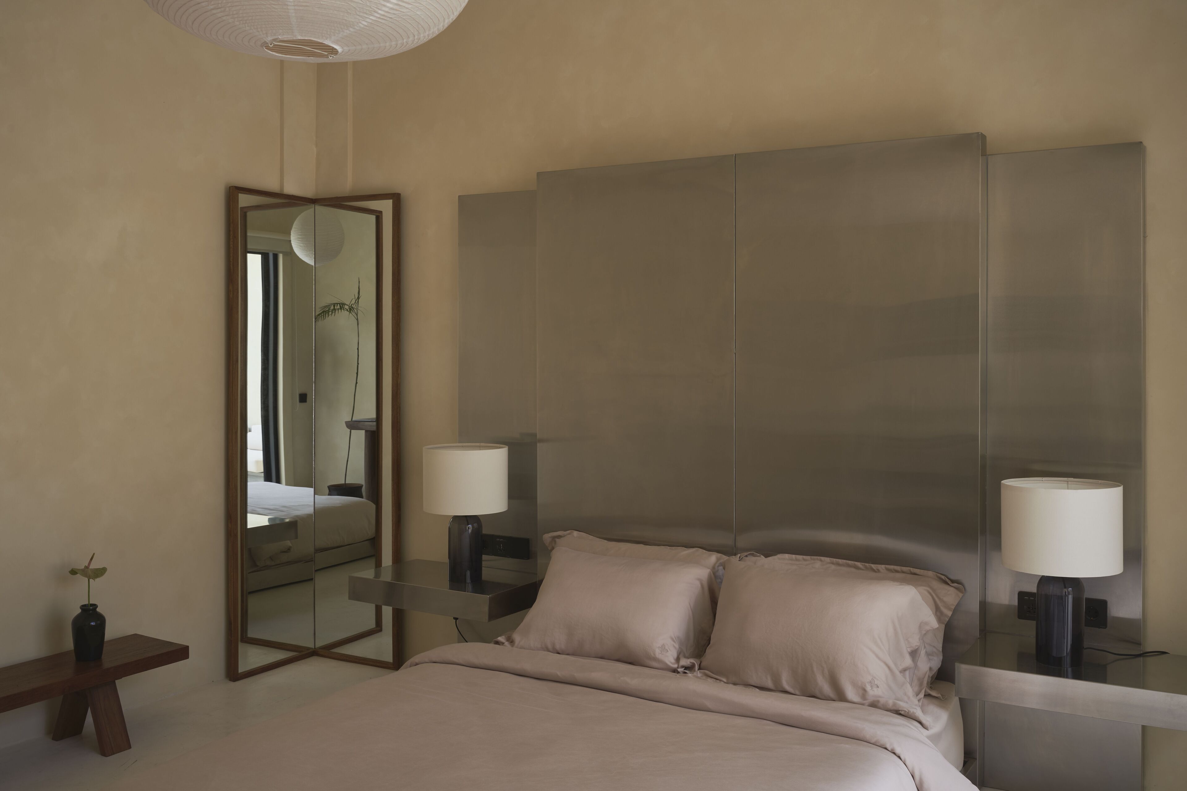 A modern hotel room with a large bed, two bedside tables with lamps, a large mirror, and a small bench with a plant.